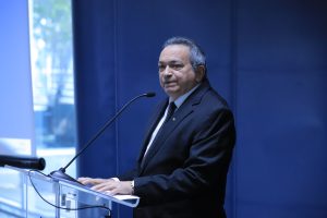 A imagem mostra um homem de meia-idade em traje formal (terno e gravata), falando em um púlpito com microfone. Ele parece estar em um ambiente profissional ou institucional, com um fundo azul. A expressão do homem é séria e focada, sugerindo que ele está fazendo um discurso ou apresentando algo. Uma janela ao lado permite a entrada de luz natural, iluminando parcialmente o ambiente.