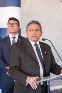 A imagem mostra dois homens em um cenário de evento ou coletiva de imprensa. O homem em primeiro plano está usando um terno escuro com risca de giz, uma camisa branca e uma gravata escura com padrões sutis. Ele está falando em um microfone, parecendo estar fazendo um discurso. Ele tem cabelo grisalho curto, rosto expressivo, e está segurando papéis ou notas na mão direita.Ao fundo, há outro homem, mais jovem, que também está vestindo um terno escuro, camisa branca e gravata com padrões pequenos. Ele usa óculos de armação grossa e parece estar prestando atenção ao que o primeiro homem está dizendo. Eles estão posicionados em frente a um fundo branco e azul, que parece ser parte de uma estrutura montada para o evento.