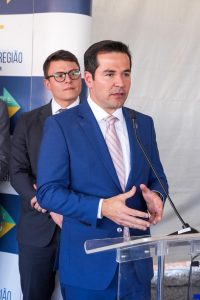 Na imagem, vemos um homem de pele clara usando um terno azul elegante e gravata listrada em tons de rosa claro. Ele está em um púlpito, falando ao microfone, com as mãos ligeiramente abertas, como se estivesse explicando ou fazendo um gesto de ênfase. Seu cabelo é escuro e bem penteado, e ele exibe uma expressão séria e atenta.Atrás dele, há outro homem mais jovem, de óculos, terno escuro e gravata, observando com atenção. Ao fundo, pode-se ver um banner que menciona o "TRT-18ª Região – Goiás", além de cores que lembram tons institucionais, sugerindo que se trata de uma inauguração oficial. 