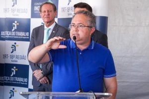 Na imagem, há um homem de meia-idade em foco, falando em um microfone durante um evento. Ele está vestindo uma camiseta polo azul com detalhes brancos nas mangas e óculos de grau. Seu cabelo é curto e grisalho, e ele gesticula com uma das mãos, como se estivesse explicando algo ou enfatizando um ponto em seu discurso.Ao fundo, está um banner com o logotipo do "TRT-18ª Região – Goiás", representando a Justiça do Trabalho. Atrás do homem que fala, há dois outros homens, um deles vestindo terno escuro e gravata verde clara, observando atentamente a fala.
