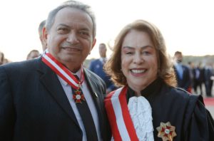 Imagem de duas pessoas sorrindo, um homem e uma mulher, ambos usando trajes formais. O homem veste um terno preto com gravata e a mulher usa uma roupa preta com detalhes brancos. Ambos estão usando medalhas e faixas vermelhas e brancas, com uma insígnia dourada no peito. Eles estão em um ambiente externo, aparentemente durante uma cerimônia ou evento formal.