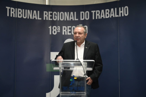 A imagem mostra um homem de meia-idade, vestido com um terno preto e sem gravata, falando em um púlpito de acrílico transparente. Ele está em frente a um painel azul que exibe as palavras "TRIBUNAL REGIONAL DO TRABALHO 18ª REGIÃO". A expressão do homem é séria e focada, indicando que ele está fazendo um discurso ou uma apresentação em um contexto oficial relacionado ao tribunal.