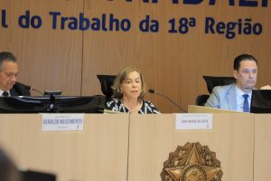 A imagem mostra uma sessão do Tribunal do Trabalho da 18ª Região. Ao centro, uma mulher fala ao microfone, acompanhada por dois outros membros do tribunal, com uma placa e brasão à frente da mesa.