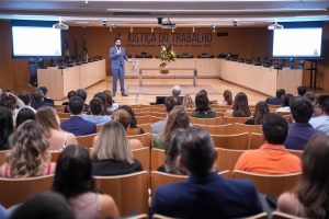 A imagem mostra um auditório com várias pessoas sentadas de frente para um palco, onde um homem de terno azul está falando ao microfone. Ao fundo, há uma mesa com cadeiras vazias e a inscrição "Justiça do Trabalho" na parede. Duas telas laterais exibem apresentações, mas o conteúdo não está visível na imagem. O público parece estar atento à apresentação.