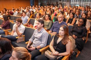A imagem mostra um auditório cheio de pessoas assistindo a uma apresentação ou palestra. Todos estão atentos ao que está acontecendo no palco. O público é composto por homens e mulheres, em sua maioria jovens adultos, sentados em cadeiras dispostas em fileiras. A atmosfera do evento é formal e organizada.
