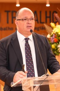 A imagem mostra um homem de óculos e terno escuro falando em um púlpito. Ele segura um microfone de lapela e está fazendo um discurso formal. Ao fundo, há uma decoração com flores e a palavra "TRABALHO" destacada em uma parede. O ambiente se trata de um evento oficial, relacionado ao âmbito jurídico e trabalhista.