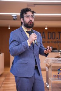 A imagem mostra um homem de barba, cabelo curto, usando óculos e terno azul. Ele segura um microfone em uma mão e um controle remoto na outra, enquanto faz uma apresentação. O homem está em um auditório, com um púlpito transparente à sua frente, que exibe o logo do TRT-18ª Região (Tribunal Regional do Trabalho de Goiás). Ao fundo, parte da inscrição "Justiça do Trabalho" é visível na parede.