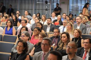 A imagem mostra um auditório cheio de pessoas assistindo a um evento. Todos estão sentados e prestando atenção, muitos usando trajes formais. Ao fundo, há uma câmera em um tripé, sugerindo que o evento está sendo gravado.
