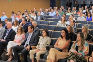 A imagem mostra um auditório com várias pessoas sentadas, assistindo atentamente a um evento. A maioria está formalmente vestida, e o ambiente é iluminado e organizado.
