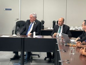 Dois homens sentados em uma mesa de negociação. Eles vestem terno preto, camisa branca e gravata azul.