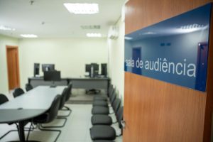 A imagem mostra a entrada de uma sala de audiência, com uma placa na porta indicando "sala de audiência". O ambiente interno parece ser simples e funcional, com uma mesa de reuniões e várias cadeiras dispostas ao redor. Ao fundo, há alguns monitores de computador sobre uma mesa. A iluminação do local é clara, provavelmente com luzes fluorescentes no teto. É um ambiente típico de um tribunal ou espaço administrativo para audiências formais.