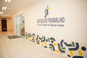 A imagem mostra um corredor claro com piso de cerâmica e uma parede decorada com o logotipo da "Justiça do Trabalho" e o nome "Vara do Trabalho de Águas Lindas". O logotipo consiste em formas geométricas estilizadas e coloridas em verde, amarelo e roxo. No lado esquerdo da imagem, há uma porta de vidro que parece dar acesso ao interior do prédio. A iluminação do local é intensa, com luzes fluorescentes no teto, e o ambiente tem um aspecto moderno e organizado, provavelmente de uma instituição pública voltada para o atendimento judicial trabalhista.