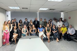 A imagem mostra um grupo de 25 pessoas em uma sala de reuniões, com algumas sentadas e outras em pé. Elas estão vestidas em trajes formais e casuais, sugerindo um encontro corporativo ou profissional. O ambiente é iluminado por luzes de teto, com uma mesa ao centro e janelas ao fundo.