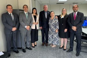 A imagem mostra um grupo de sete pessoas em pé, lado a lado, posando para uma foto em um ambiente de escritório. Todos estão vestidos formalmente, com trajes sociais como ternos e vestidos. O grupo parece estar em um ambiente profissional, provavelmente após uma reunião ou evento corporativo. O fundo revela uma sala típica de escritório, com móveis e computadores ao redor.