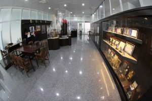 A imagem mostra um ambiente interno moderno com piso de granito, móveis de madeira e uma grande vitrine à direita, exibindo vários objetos e documentos emoldurados. No fundo, há uma área com mesas e cadeiras, sugerindo que o espaço é um local de exposição com itens históricos e culturais em exibição.