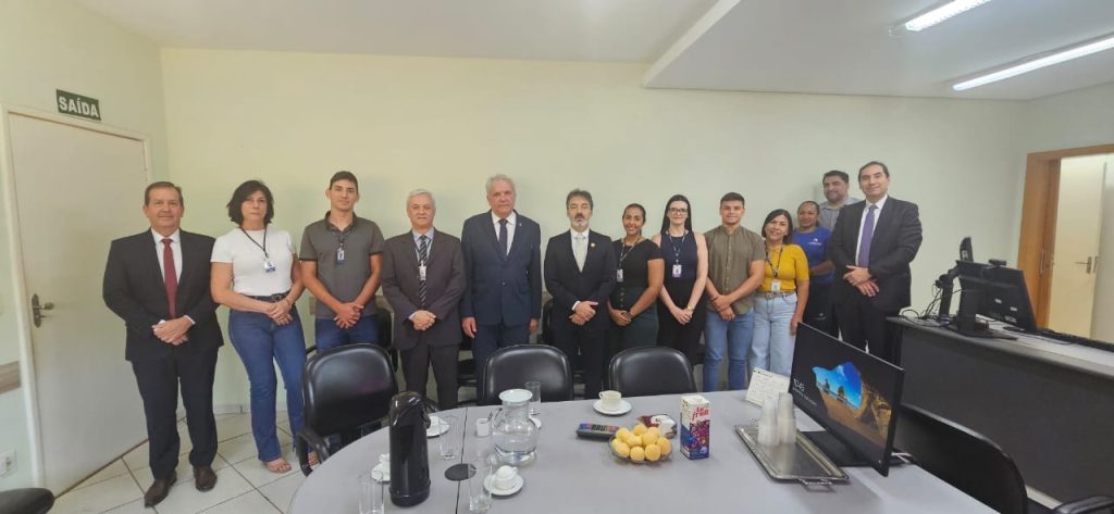 A imagem mostra um grupo de pessoas posando em frente a uma mesa de reunião. Há uma mistura de homens e mulheres, com alguns vestindo trajes formais (como ternos) e outros com roupas mais casuais. No centro, há um homem de terno que parece ser uma figura de destaque, cercado por outras pessoas que estão em pé ao redor da mesa. Na mesa, há copos, garrafas e alguns alimentos, sugerindo que se trata de uma reunião ou evento formal com uma pausa para café.