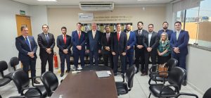 A imagem mostra um grupo de pessoas em uma sala de reuniões formal. A maioria dos presentes são homens vestidos com ternos e gravatas, além de uma mulher à direita, vestida de maneira formal. No centro da foto, há um homem de terno claro, que parece ser uma figura de destaque. Todos estão alinhados em frente a uma mesa de reunião, em um ambiente que parece ser institucional. Há cadeiras ao redor da mesa e uma janela à direita, permitindo a entrada de luz natural.