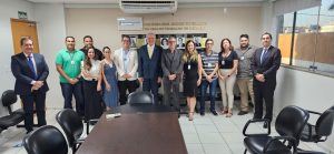 A imagem mostra um grupo de pessoas posando para uma foto em um ambiente formal, provavelmente uma sala de reuniões. No centro, há um homem de terno que parece ser uma figura de autoridade, cercado por outras pessoas, incluindo homens e mulheres. Eles estão alinhados em frente a uma mesa de reunião, e todos estão olhando para a câmera. 