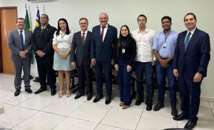 A imagem mostra um grupo de pessoas em pé, posando para a foto em um ambiente fechado. O grupo é composto por homens e mulheres, e ao fundo é possível ver bandeiras do Brasil e de Goiás, indicando que o encontro tem um contexto oficial ou institucional.
