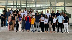 A imagem mostra um grupo de pessoas posando para uma foto em frente a um prédio com janelas grandes. As pessoas estão em trajes casuais e formais, posicionadas lado a lado, sorrindo para a câmera.