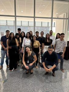 A imagem mostra um grupo de pessoas posando para uma foto em um ambiente interno com janelas grandes ao fundo. Eles estão sorrindo para a câmera e usam trajes casuais e sociais. Duas pessoas estão agachadas na frente, enquanto as demais estão em pé, formando uma pose descontraída.