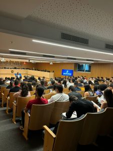 A imagem mostra um auditório cheio de pessoas sentadas. A maioria está virada para a frente, onde há um apresentador e uma tela azul exibindo o logo da Justiça do Trabalho. O ambiente é bem iluminado e organizado, com cadeiras de madeira e um clima formal.