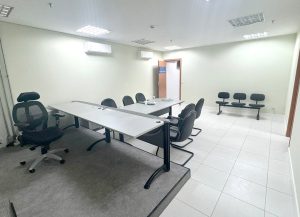 A imagem mostra uma sala de audiências. No centro, há uma mesa longa e branca com várias cadeiras de escritório pretas ao redor. Uma das cadeiras está em uma pequena plataforma elevada. No fundo, à direita, há mais algumas cadeiras de espera encostadas na parede. O ambiente é iluminado por luzes de teto, com piso de azulejo claro, paredes de cor neutra e um ar-condicionado montado na parede.