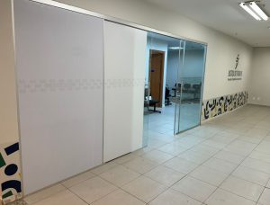 A imagem mostra o interior de um ambiente limpo e moderno. Há uma parede de vidro transparente que divide o espaço, e no fundo, é possível ver cadeiras dispostas em uma sala de espera. À direita, na parede, há a logo da Justiça do Trabalho. A decoração é minimalista, com piso claro e uma faixa decorativa em cores vivas na parede à direita. 