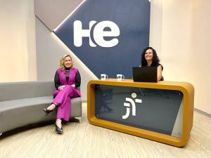 A imagem mostra duas mulheres em um ambiente de estúdio. Uma está sentada em um sofá cinza, vestindo um conjunto rosa, e a outra está atrás de uma bancada, usando roupas pretas e com um laptop à sua frente. A bancada possui um logotipo da Justiça do Trabalho. No fundo, há um grande painel com as letras "HE". 