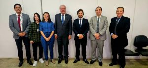 A imagem mostra um grupo de sete pessoas alinhadas, todas de pé, com fundo branco. Algumas usam trajes formais, enquanto outras estão vestidas de forma mais casual.