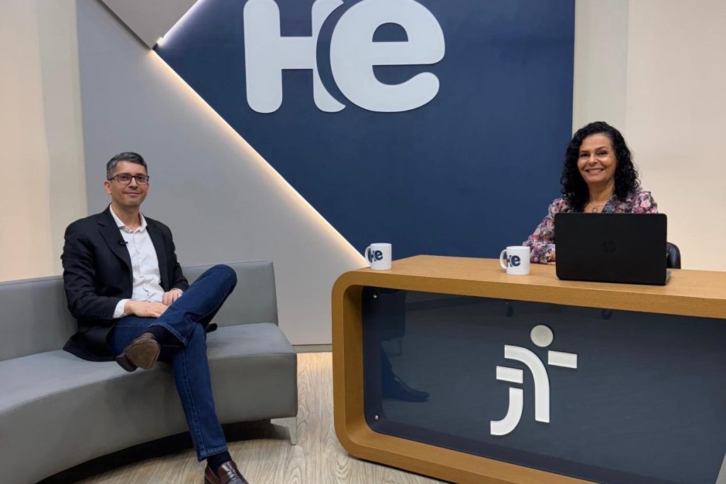 Estúdio de tv com uma apresentadora e um entrevistado. Ele está sentado no sofá e ela numa bancada.