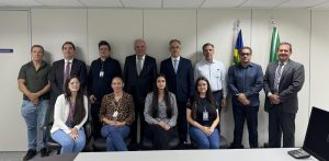 A imagem mostra um grupo de 12 pessoas em um ambiente corporativo, com sete em pé e cinco sentadas, posando para uma foto formal. Ao fundo, estão as bandeiras do Brasil e outra possivelmente institucional, indicando um evento oficial ou reunião.