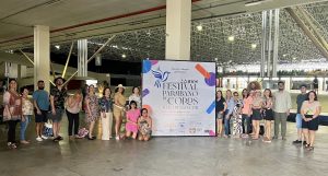 A imagem mostra um grupo de pessoas alinhadas em um espaço interno amplo, com estrutura metálica no teto. Elas estão ao lado de um painel que anuncia o "22º Festival Paraibano de Coros", realizado em João Pessoa, PB, de 18 a 21 de novembro de 2021, com homenagem ao maestro Eduardo Lakschevitz. O local parece ser um centro de convenções ou um espaço multiuso. O grupo aparenta ser formado por participantes ou organizadores do evento.