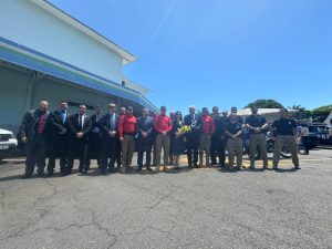 A imagem mostra um grupo de pessoas posando para uma foto ao ar livre, sob um céu claro. Há uma linha de indivíduos, alguns em trajes formais, como terno e gravata, e outros vestindo uniformes ou roupas mais casuais. O cenário parece ser a parte externa de um prédio, com um toldo azul visível à esquerda. É uma foto de grupo, relacionada a um evento institucional.