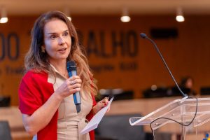 A imagem mostra uma mulher segurando um microfone azul em uma das mãos e um documento na outra. Ela está em pé ao lado de um púlpito transparente, aparentando estar discursando ou apresentando algo. Ela veste uma roupa formal clara com um casaco vermelho e parece estar em um ambiente corporativo ou institucional. O fundo desfocado revela um painel com parte de um texto visível, sugerindo o contexto de uma conferência ou evento oficial.