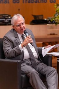 A imagem mostra o desembargador Platon Filho. Ele tem cabelos grisalhos, usa terno cinza e gravata e está sentado em uma poltrona enquanto fala ao microfone. Ele segura papéis na outra mão, sugerindo que está participando de um evento formal ou painel de discussão. Ao fundo, é possível ver um ambiente corporativo ou institucional.