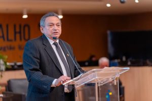 A imagem mostra o desembargador Geraldo Nascimento. Ele é um homem de terno escuro, camisa branca e gravata listrada, falando em um púlpito transparente. Ele está equipado com um microfone de lapela e parece estar fazendo uma apresentação ou discurso em um ambiente formal. O fundo da imagem mostra uma parede com escrita parcialmente visível e um ambiente interno bem iluminado, sugerindo um contexto corporativo ou institucional.