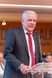 A imagem mostra o desembargador Eugênio Cesário em pé, de frente ao púlpito, fazendo seu discurso. Ele é um homem alto, de cabelos grisalhos, usa terno preto, camisa branca e gravata vermelha.