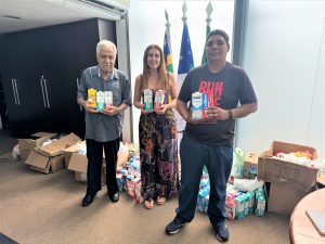 A imagem mostra três pessoas segurando alimentos e produtos de higiene, com caixas ao fundo, sugerindo um evento de doação em ambiente institucional.