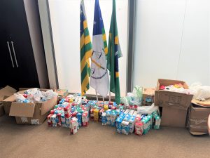 A imagem mostra caixas e embalagens de leite organizadas ao lado de bandeiras institucionais, indicando uma entrega ou arrecadação de alimentos em um evento formal.