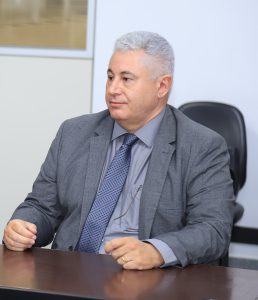 Homem de cabelos grisalhos, terno cinza e gravata azul, sentado à mesa em ambiente formal, com expressão atenta.