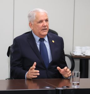 Homem de cabelos grisalhos, terno escuro e gravata azul, sentado e gesticulando em uma reunião formal.