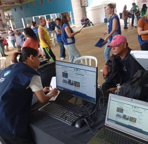 Uma mulher com uniforme azul escuro está inclinada sobre uma mesa, preenchendo um papel, enquanto um homem com boné vermelho e jaqueta preta está sentado em frente a ela. A mesa contém computadores e laptops conectados, exibindo uma página de um site. No fundo, há várias pessoas caminhando, algumas com pranchetas, em um espaço coberto com divisórias e placas informativas.