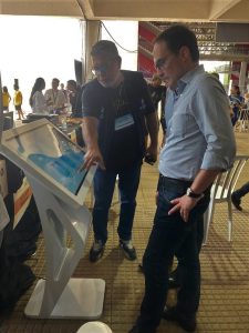 Um homem usando óculos e vestindo um colete azul escuro aponta para um monitor touchscreen em um suporte, enquanto outro homem, de camisa social azul-clara e calça jeans, observa atentamente. O local parece um ambiente de evento, com várias pessoas ao fundo interagindo e transitando. Há também um balcão com itens visíveis como garrafas e alimentos.