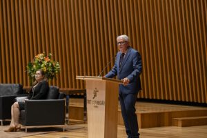 Sob o palco de um auditório, um homem branco de terno fala ao microfone.