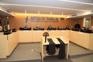 Grupo de pessoas em uma reunião, eles estão sentados em uma bancada de frete ao público. Atrás deles há um letreiro escrito "Justiça do Trabalho, Tribunal Regional do Trabalho da décima oitava região-GO.".