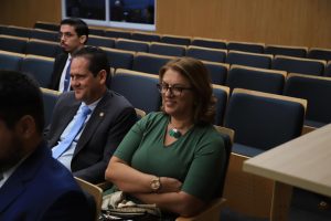 Mulher sentada em um auditório ao lado de um homem usando terno, ela está sorrindo com os braços cruzados e vestindo uma blusa verde, usa um colar com detalhes de folhas e cabelos na altura do ombro.