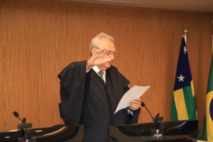 Homem usando roupas formais e com a mão direita levantada fazendo sinal de juramento.