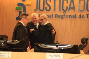 Três homens em pé se abraçando atrás de uma bancada, todos estão usando toga e há atrás deles um letreiro escrito "Justiça do Trabalho, Tribunal Regional do Trabalho da décima oitava região-GO".