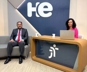 Um homem sentado em uma poltrona e uma mulher numa bancada. Eles olham para a câmara. Ao fundo, a logomarca do programa hora extra. Embaixo da bancada, a logomarca da Justiça do Trabalho